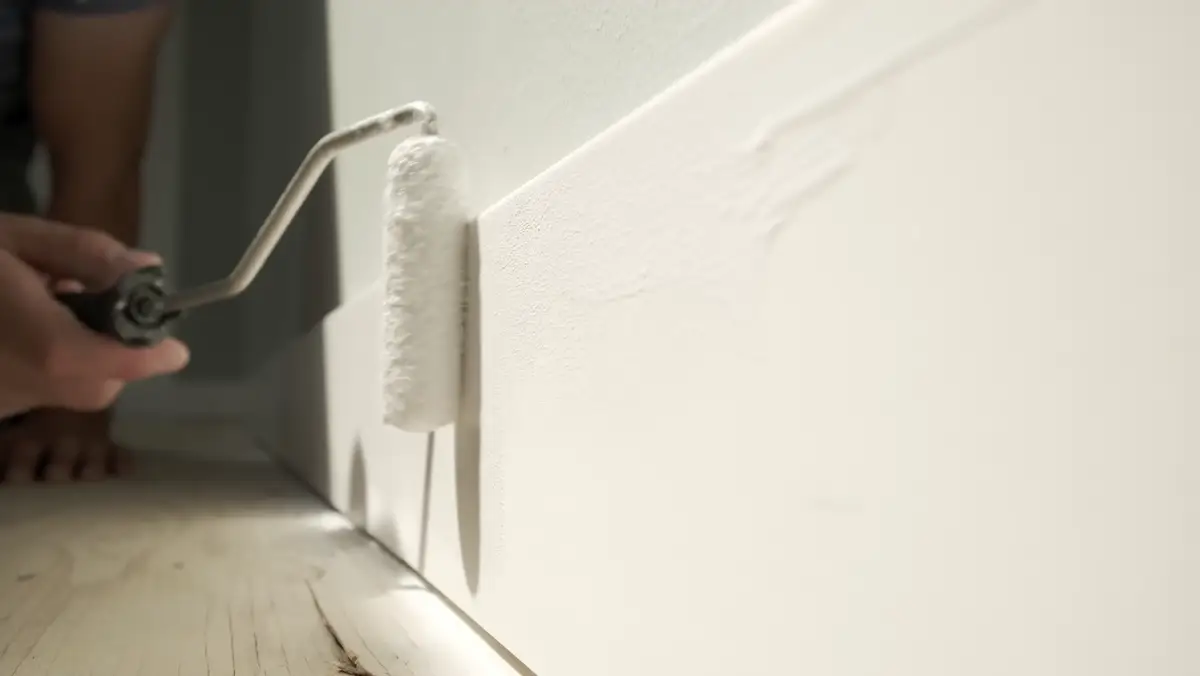 Roller applying white paint along a baseboard